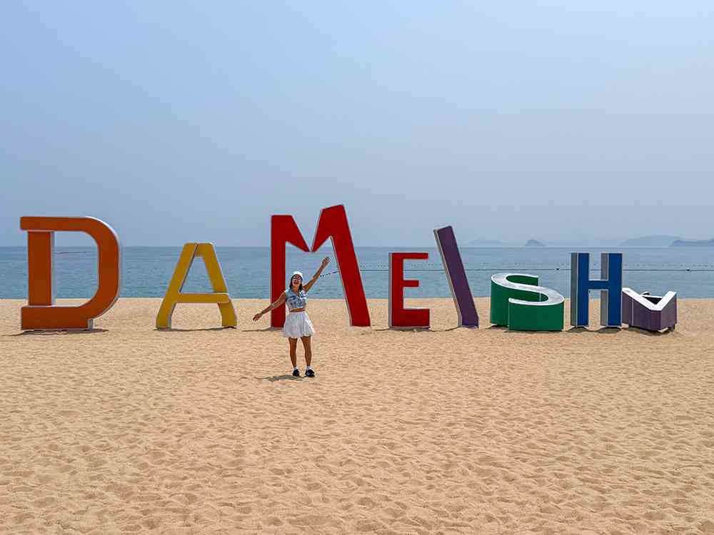Letras gigantes de colores del cartel "DAMEISHA" en la playa de Dameisha (大梅沙), Shenzhen, con la arena dorada, el mar azul y las montañas verdes de fondo.