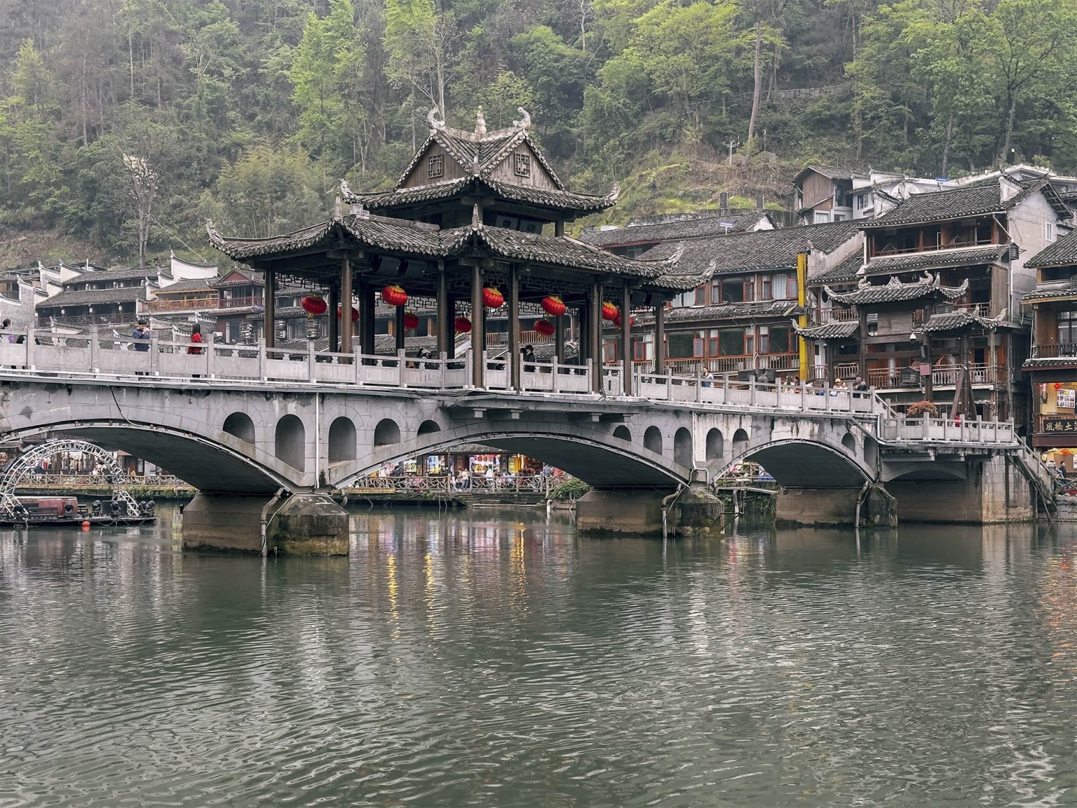 Puente del Viento y la Lluvia (Fengyuqiao) cubierto con tejado de madera y aleros curvos, cruzando el río Tuojiang en Fenghuang. El puente de madera tallada con bancos para sentarse refleja la arquitectura tradicional Miao junto a las casas colgantes del pueblo del Fénix