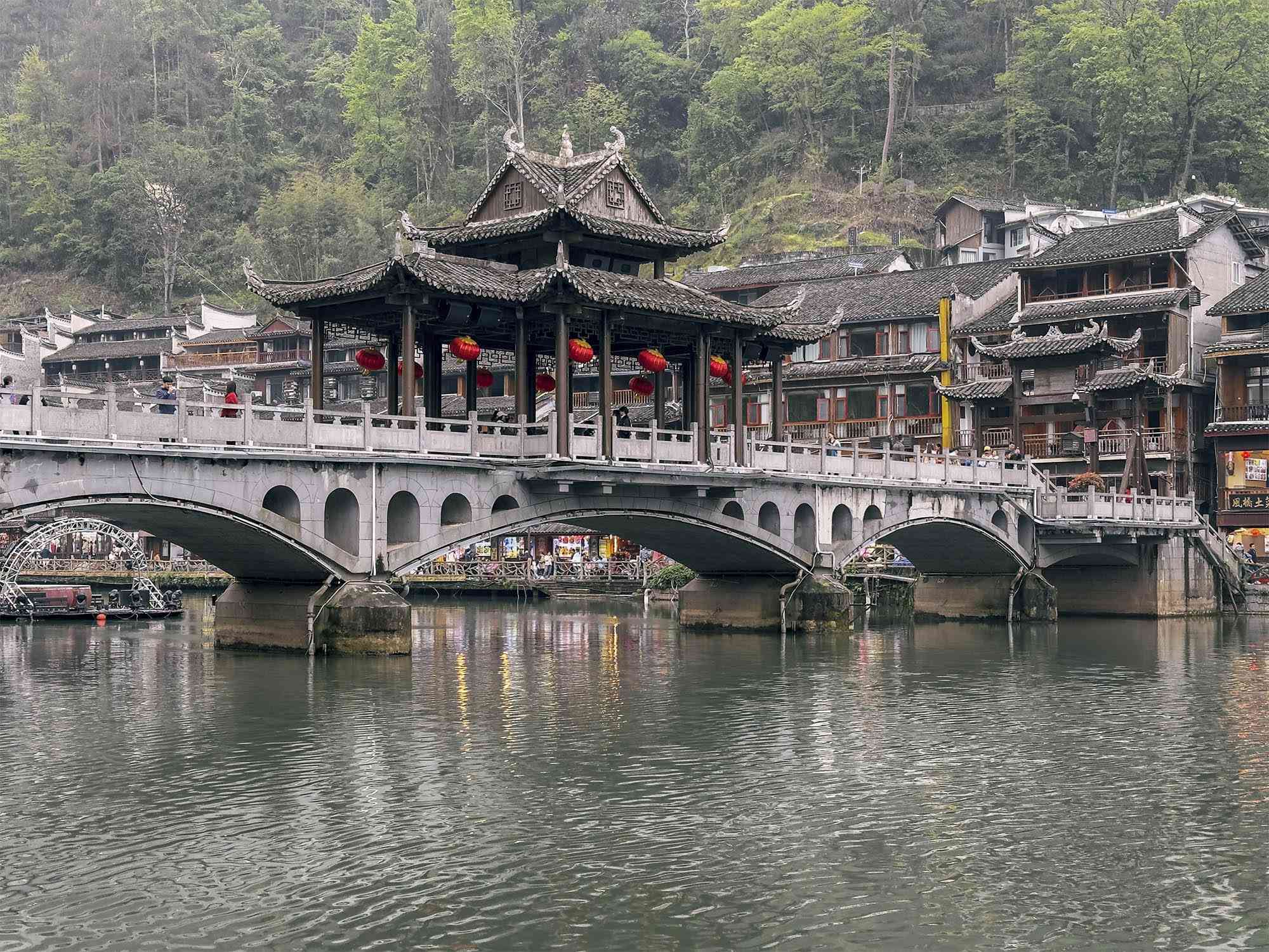 Puente del Viento y la Lluvia (Fengyuqiao) cubierto con tejado de madera y aleros curvos, cruzando el río Tuojiang en Fenghuang. El puente de madera tallada con bancos para sentarse refleja la arquitectura tradicional Miao junto a las casas colgantes del pueblo del Fénix