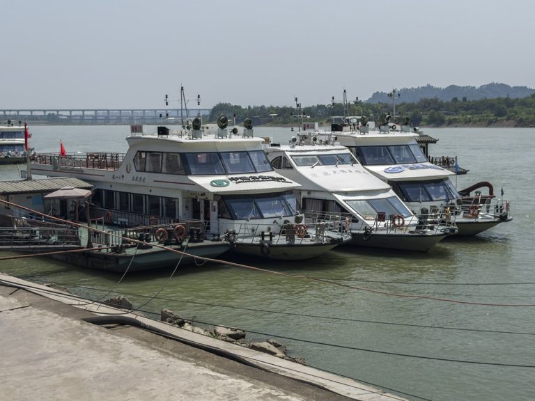 Vista del puerto de Leshan con varios barcos turísticos amarrados en la orilla, flotando tranquilamente en el río, con la silueta del Buda de Leshan y las montañas verdes de fondo bajo una luz suave de atardecer.