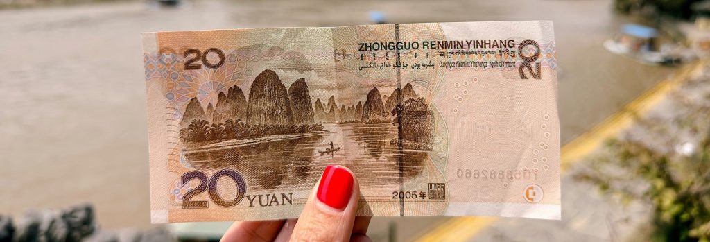 Billete de 20 yuanes chinos (RMB) sujetado frente al paisaje real de las montañas de Xingping, en la región de Guilin, China. El dibujo del billete se alinea con el paisaje de fondo, mostrando las emblemáticas montañas kársticas y el río Li. Foto de portada para la guía sobre cómo pagar en China en 2026.