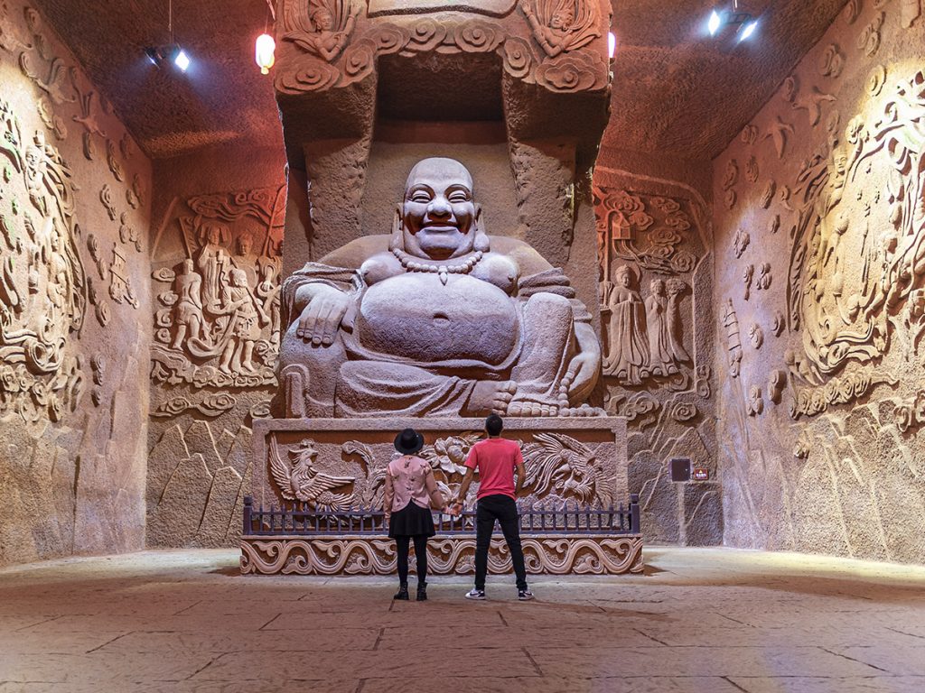 Dos personas mirando con admiración a una estatua de Buda sonriente en una sala de la Cueva de los 10.000 Budas, con las paredes de roca completamente cubiertas de cientos de pequeños grabados y relieves tallados a mano.