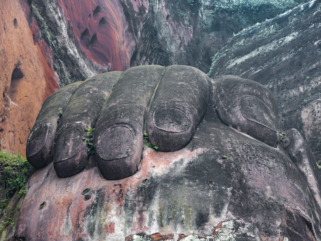 Primer plano de la mano derecha del Gran Buda de Leshan, con los dedos gruesos y la palma tallada en piedra, apoyada sobre la rodilla, mostrando la textura y la escala colosal de la escultura.