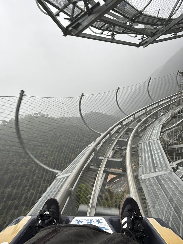 Marta y Javi descendiendo en un trineo monoplaza sobre raíles por el circuito de montaña de Gulongxia en Qingyuan, Guangdong, China. El trazado tiene 3,68 kilómetros de longitud, 260 metros de desnivel y 28 curvas, incluyendo varias en horquilla. Una experiencia de velocidad y adrenalina entre bosques y montañas.