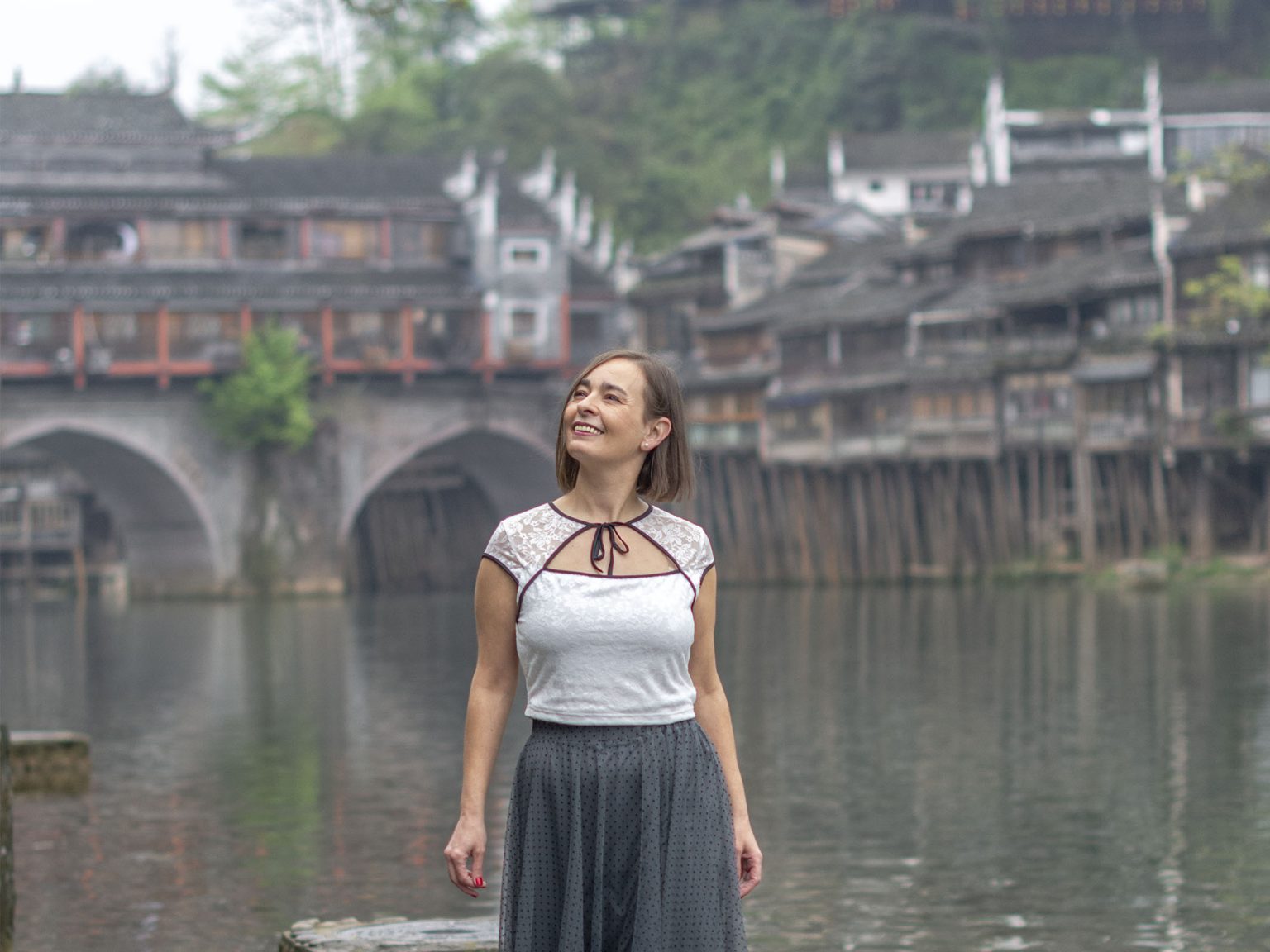 Mujer paseando por el sendero junto al río Tuojiang en Fenghuang, con las casas colgantes de madera sobre pilotes, farolillos rojos y el Puente del Arcoíris al fondo en el pueblo del Fénix