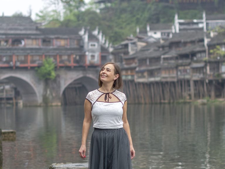 Mujer paseando por el sendero junto al río Tuojiang en Fenghuang, con las casas colgantes de madera sobre pilotes, farolillos rojos y el Puente del Arcoíris al fondo en el pueblo del Fénix