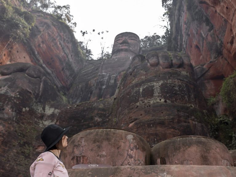 Marta de pie junto a los pies del Gran Buda de Leshan, mirando hacia arriba con asombro, con el coloso de piedra de 71 metros elevándose majestuoso frente a ella, bajo un cielo despejado.