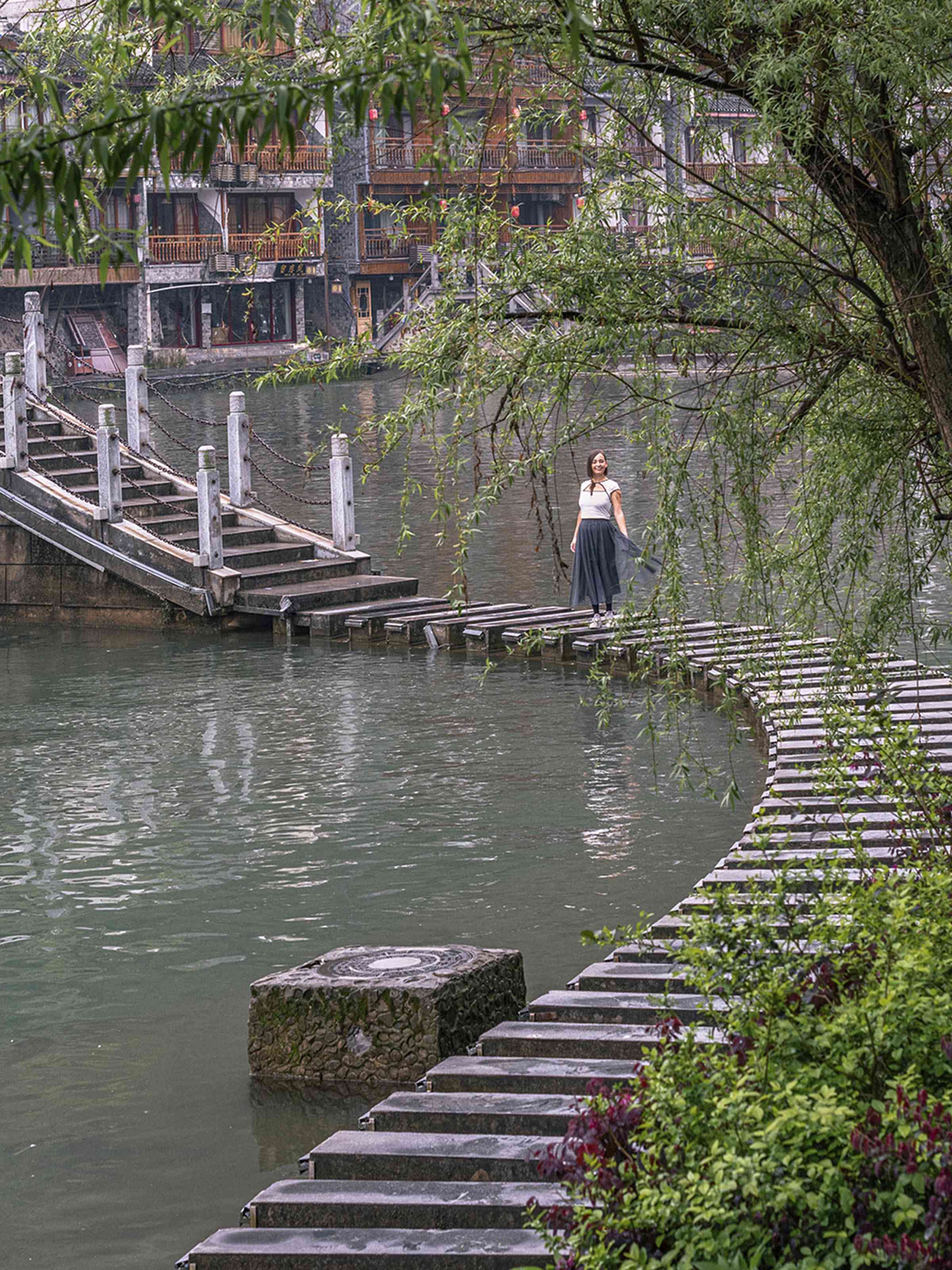 Marta caminando por el Puente de la Luna (Yueqiao) en Fenghuang, Hunan. El original puente arqueado con pasarelas de piedra exteriores cruza el río Tuojiang, con una luz suave de amanecer y las casas colgantes de madera del pueblo del Fénix al fondo
