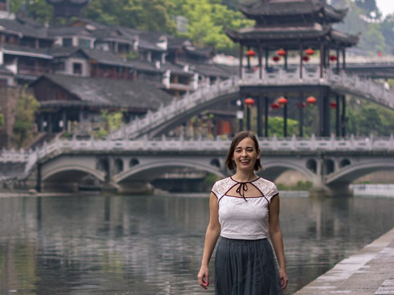 Mujer caminando por el paseo junto al Puente de Nieve (Xueqiao) sobre el río Tuojiang en Fenghuang. El puente de madera de dos niveles con pabellón central diseñado por Huang Yongyu se alza detrás, con las casas colgantes y los árboles de fondo
