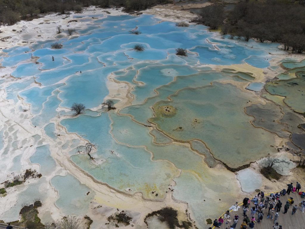 Vista aérea de las piscinas de travertino de Huanglong en Sichuan, con formaciones de color turquesa y blanco que se extienden como terrazas escalonadas por la ladera de la montaña, rodeadas de bosque y picos nevados al fondo.