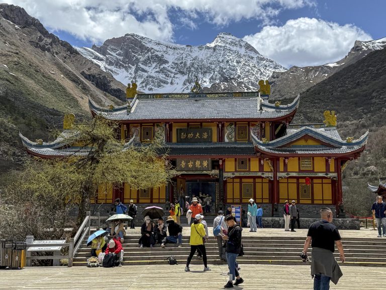 El Templo de Huanglong (黄龙古寺), de paredes rojas y techos oscuros, se alza a 3.558 metros de altitud con los picos nevados de la cordillera Minshan, incluyendo el Xuebaoding (5.588 m), como imponente telón de fondo bajo un cielo azul.