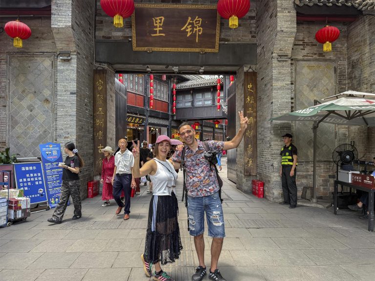 Dos viajeros posando en la entrada de Jinli Street (锦里古街) en Chengdu, con el arco de entrada de madera tallada con los caracteres "锦里" y farolillos rojos chinos de fondo, bajo un cielo de atardecer.
