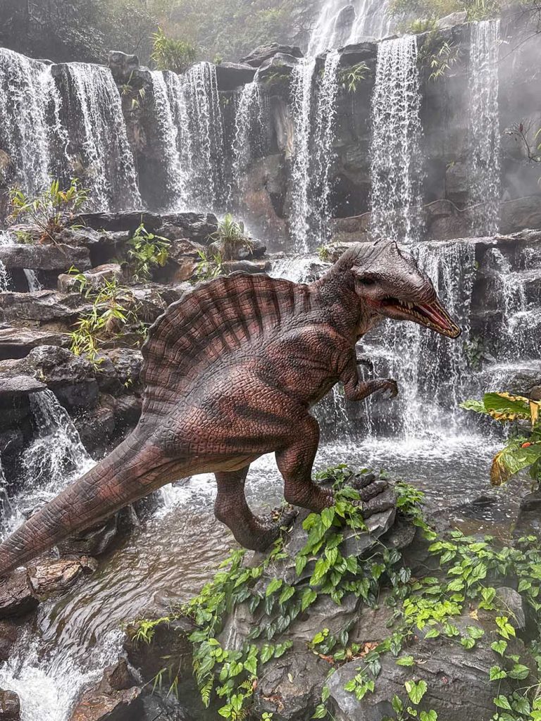 Primer plano de un dinosaurio animatrónico con la cascada de Gulong Gorge al fondo, Qingyuan, China