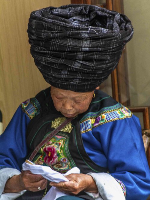 Mujer mayor de la etnia Miao con ropaje tradicional oscuro y gran tocado de plata en la cabeza, sentada bordando a mano con aguja e hilo en un taller de Fenghuang, Hunan. La artesana Miao luce una corona de plata con motivos de fénix, transmitiendo las técnicas de bordado tradicional que las niñas aprenden desde la infancia
