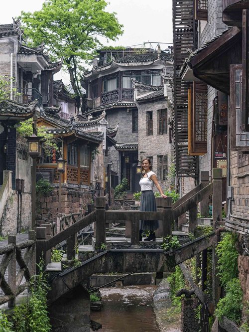 Callejón empedrado de Fenghuang con casas tradicionales de madera de las dinastías Ming y Qing, farolillos rojos colgando, tejados curvados de tejas negras y pequeños comercios locales. La calle antigua, con su atmósfera de pueblo congelado en el tiempo, es uno de los rincones más auténticos del casco histórico