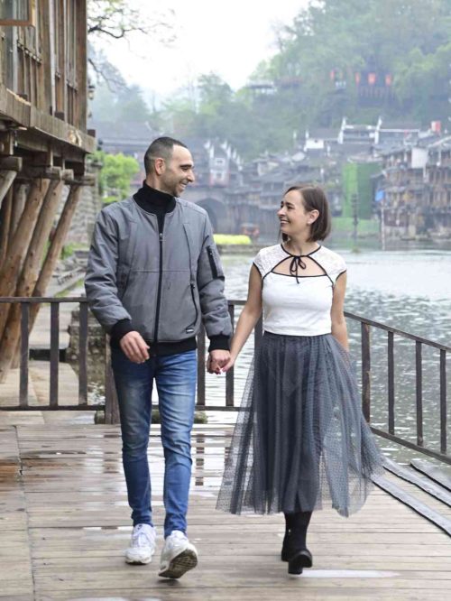Mujer con traje tradicional Miao de color rojo brillante y joyas de plata posando junto al río Tuojiang en Fenghuang. La modelo lleva el elaborado tocado de plata y el collar característico de la etnia Miao, con las casas colgantes y el Puente del Arcoíris al fondo