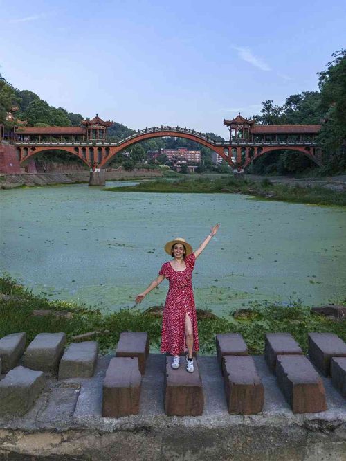 Fotografía tomada con drone que muestra a la viajera Marta, vestida con un vestido rojo de lunares y sombrero de paja, posando alegremente sobre un antiguo sendero de bloques de piedra a orillas del río. En el plano medio, el majestuoso Puente Haoshan (con su arquitectura roja de pabellones tradicionales) cruza el río cubierto de algas verdes, rodeado de un exuberante paisaje montañoso bajo un cielo azul despejado.
