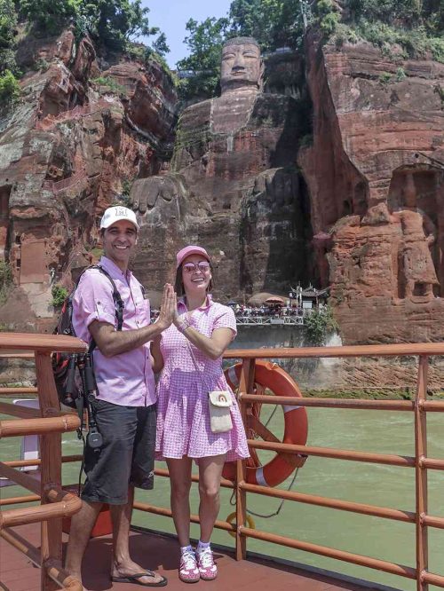 Nosotros dos sonriendo en la cubierta de un barco turístico navegando por el río, con el colosal Gran Buda de Leshan tallado en la montaña de fondo, rodeado de vegetación y bajo un cielo despejado.