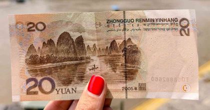 Billete de 20 yuanes chinos (RMB) sujetado frente al paisaje real de las montañas de Xingping, en la región de Guilin, China. El dibujo del billete se alinea con el paisaje de fondo, mostrando las emblemáticas montañas kársticas y el río Li. Foto de portada para la guía sobre cómo pagar en China en 2026.