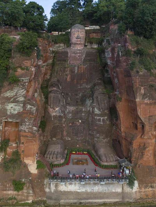 Vista aérea con drone del colosal Buda tallado en la ladera de una montaña rocosa, rodeado de vegetación y pequeños templos tradicionales chinos a sus pies, bajo un cielo azul despejado.