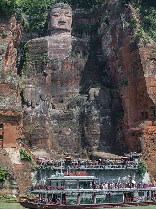Imagen aérea de drone del Gran Buda de Leshan de 71 metros de altura visto a cuerpo completo, tallado en el acantilado junto a la confluencia de los ríos Min, Dadu y Qingyi, con un barco turístico navegando frente a la estatua