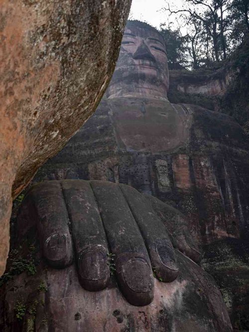 Vista desde abajo del colosal Buda de Leshan tallado en la montaña, con una de sus enormes manos apoyada sobre la rodilla en primer plano, destacando el tamaño gigantesco de la escultura frente al cielo azul.