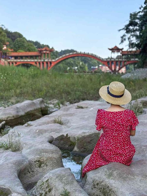 Vista del Puente Haoshan (濠上大桥) en Leshan, un puente peatonal de estilo chino con arcos, pabellones y barandillas de madera, reflejado simétricamente en el río Mahao, con el fondo de la vegetación de la montaña y el cielo despejado.