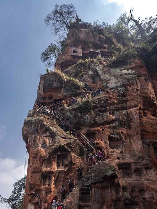 Vista de las empinadas escaleras de piedra de los Nueve Recodos, talladas junto al Buda de Leshan, con pasamanos de hierro, escalones desgastados y el río y la vegetación de fondo en un día soleado.