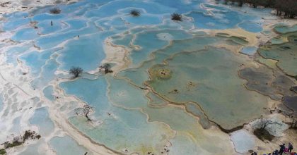 Vista aérea de las piscinas de travertino de Huanglong en Sichuan, con formaciones de color turquesa y blanco que se extienden como terrazas escalonadas por la ladera de la montaña, rodeadas de bosque y picos nevados al fondo.