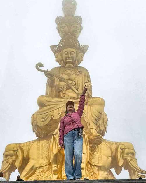 Marta de pie en la cima del Monte Emei, con los brazos abiertos y una sonrisa, rodeada de picos montañosos, niebla o mar de nubes y templos tradicionales chinos al fondo bajo un cielo azul.