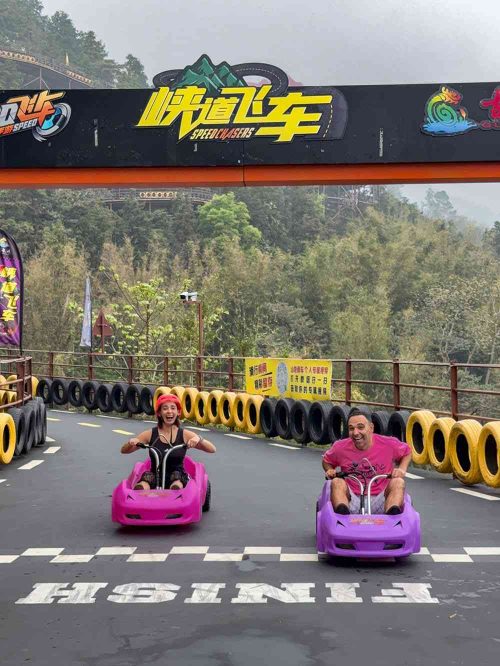 Marta y Javi descendiendo por el circuito de Gulongxia en Guangdong, China, montados en dos coches de carreras estilo Mario Kart. El trazado serpentea entre montañas y bosques con curvas cerradas y vistas espectaculares. Una de las experiencias más divertidas en los parques de aventura de China.