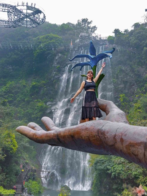 Marta subida en la Mano de Buda (通天佛手) de Gulongxia en Qingyuan, Guangdong, China. La impresionante mano gigante de 20 metros de largo y 16 metros de alto se extiende desde el acantilado como un trono de loto, y detrás de ella se ve una espectacular cascada rodeada de montañas verdes. Una de las fotos más icónicas en los parques de aventura del sur de China.